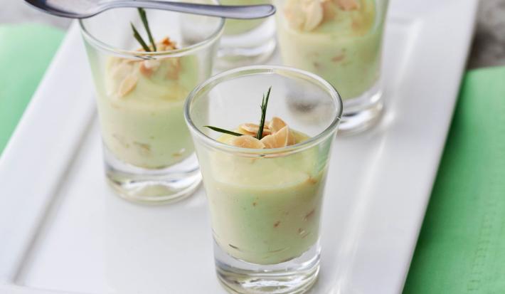 Fotografia em tons de verde em uma bancada de madeira com guardanapo verde, um prato retangular branco raso ao centro com quatro copinhos de vidro com o brigadeiro de capim-santo e lascas de amêndoas dentro deles.