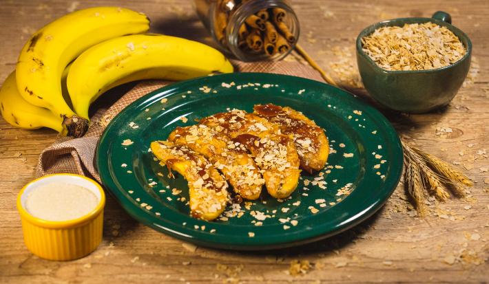 Foto da receita de banana dourada com Neston, sobre um prato verde, decorada com Neston 3 Cereais, sobre uma bancada de madeira decorada com bananas, trigo e alguns potes com açúcar e cereal dentro
