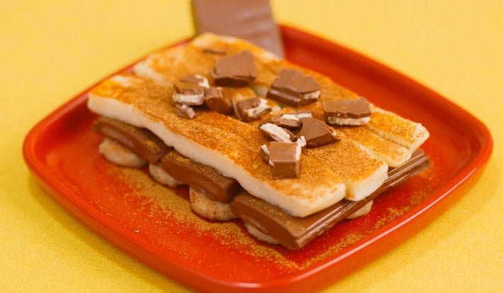 Sobremesa em prato vermelho com banana, creme claro, chocolate recheado e canela por cima.