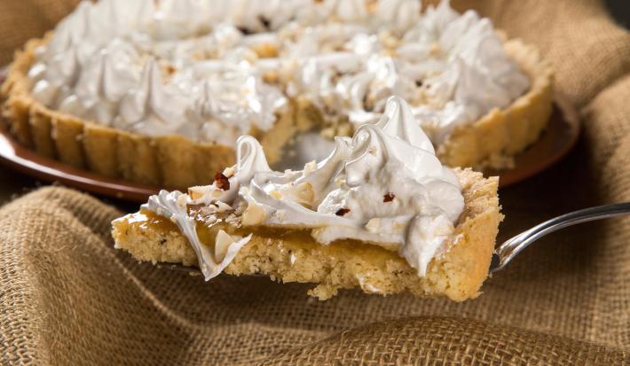 Fotografia de uma espátula segurando uma fatia de torta de cupuaçu e castanha, com merengue por cima. Ao fundo, a mesma torta, inteira sobre apoio redondo na cor marrom.