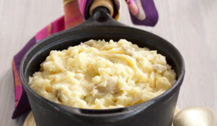 Foto de uma panela pequena na cor preta, cheia de purê de batatas com alho e uma colher prata próximo da panela
