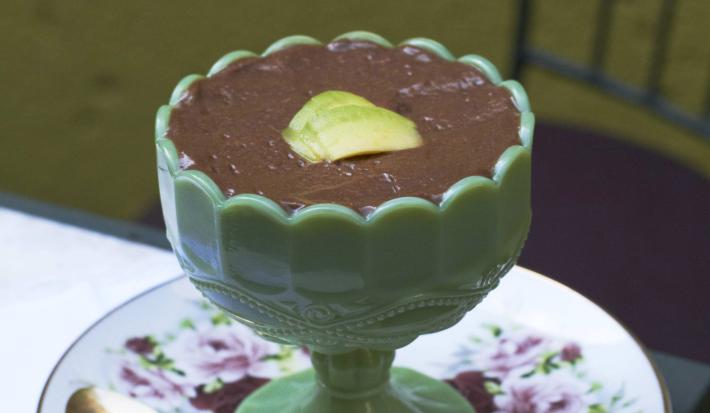 Fotografia em tons de verde em uma mesa de madeira com uma toalha branca de renda, um prato redondo branco com flores roxas e folhas verdes, uma taça verde com a mousse de abacate com chocolate dentro dela.