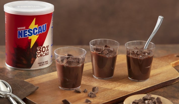 Fotografia de uma tábua retangular de madeira, pequena, com três potinhos de acrílico com o brigadeiro cremoso de colher de nescau. Ao lado tem a embalagem de NESCAU Extra Cacau 30%.