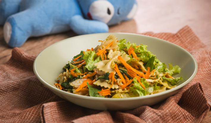 Fotografia em tons de verde e marrom em uma bancada de madeira, um prato redondo fundo verde com a salada de macarrão, frango, legumes e molho pesto dentro. Ao fundo, um ursinho azul deitado.