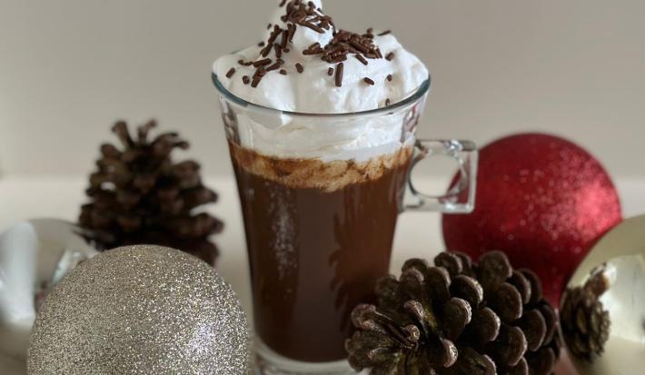 Fotografia de uma xícara de vidro longa com uma receita de espresso cremoso com chocolate, chantilly por cima e um pouco de granulado decorando. A xícara está sobre uma mesa branca, e rodeando da xícara tem alguns enfeites de natal.