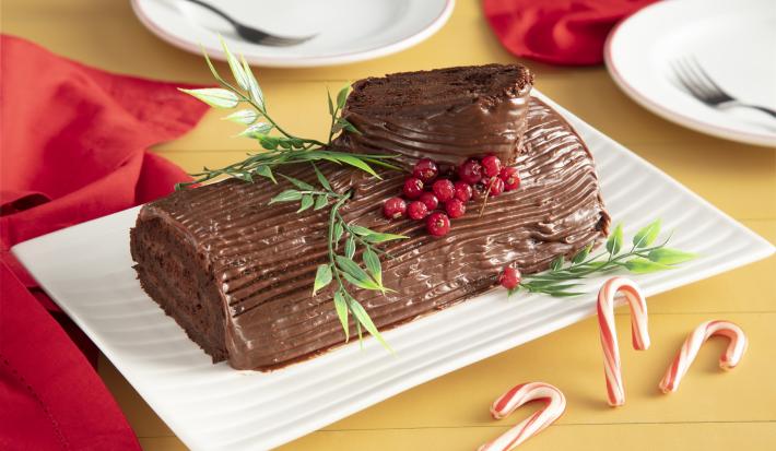 Fotografia em tons de vermelho em uma bancada de madeira amarela, um pano vermelho ao lado e ao centro, um recipiente retangular branco com o rocambole de chocolate em cima com decoração natalina.