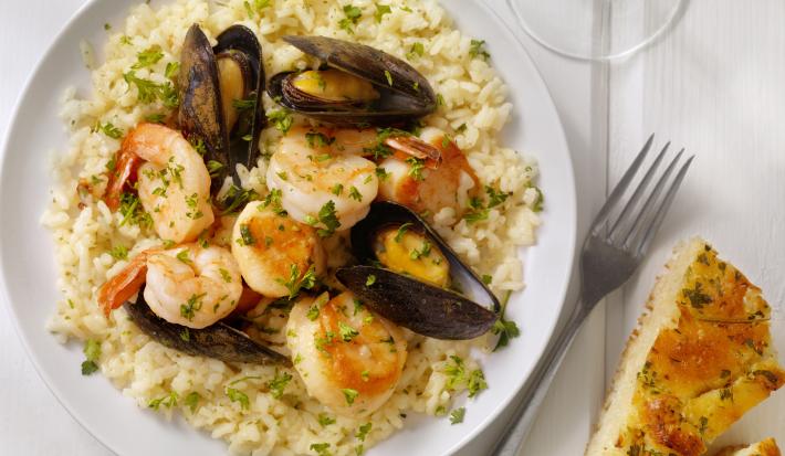 Fotografia em tons de branco e bege de uma bancada branca vista de cima. Contém um prato redondo branco com risoto com mariscos e ao lado um garfo para servir.