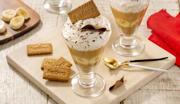 Fotografia em tons de marrom claro com duas taças de vidro ao centro. Dentro de cada taça existe uma receita de banoffee com camadas de biscoito TOSTINES, banana, doce de leite e chantilly.