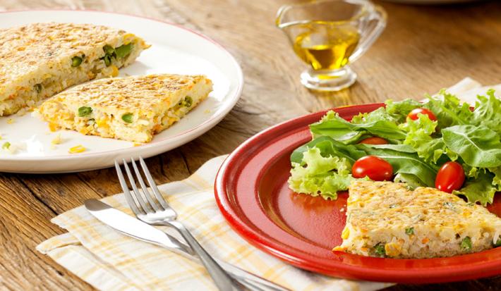 Fotografia em tons de vermelho e amarelo em uma bancada de madeira com um prato branco raso redondo ao fundo com a tortilla de arroz e ao lado, um prato vermelho com uma fatia da tortilla e salada de alface e tomate cereja.