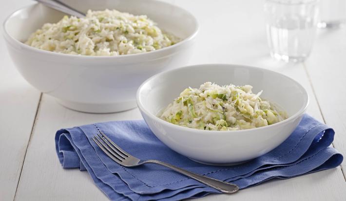 Fotografia em tons de branco e azul de uma bancada de madeira, sobre ela um paninho azul com um recipiente redondo com o arroz. Ao fundo um recipiente redondo com arroz, uma colher e um copo de vidro com água.