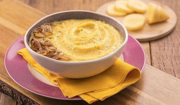 Foto em tons de amarelo da receita de creme de mandioquinha com cebola dourada servida em um bowl de porcelana branco sobre pano amarelo em cima de um prato com borda cor de rosa. Esses elementos sobre uma tábua de madeira e ao fundo mandioquinha cortada