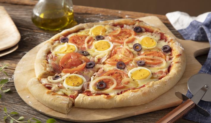 Imagem de uma pizza portuguesa redonda com queijo, tomate, cebola, ovo, azeitona preta sobre uma tábua de madeira.