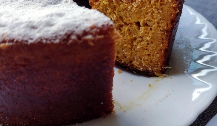 Fotografia em tons de bege em uma bancada com toalha cinza, um prato branco redondo raso, um bolo de milho cortado ao meio com açúcar polvilhado em cima.