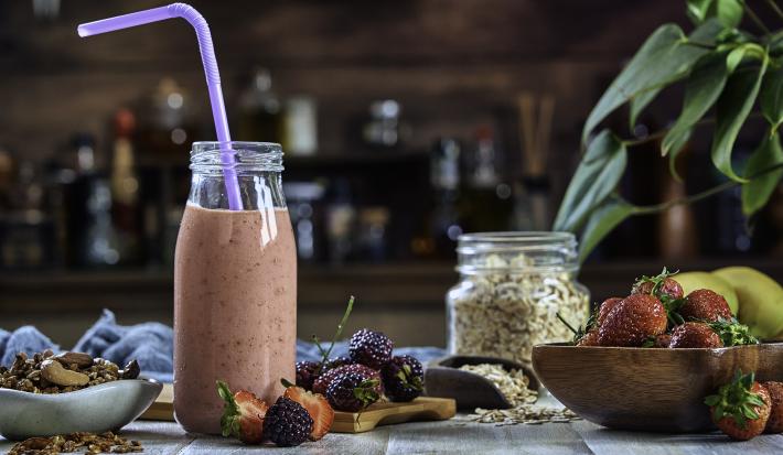 Fotografia de uma bebida densa de morango com leite MOÇA e neston que está em uma garrafa de vidro decorativa, com um canudo roxo e frutas no fundo.