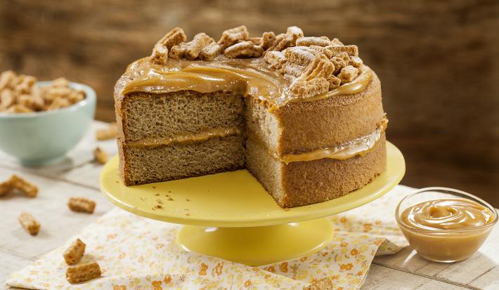 Fotografia em tons de amarelo em uma mesa de madeira com um pano bege com flores laranja e amarela, um suporte amarelo com o bolo de Moça Churros. Ao lado, um potinho com doce de leite. Ao fundo, um potinho azul com churros pequenos.