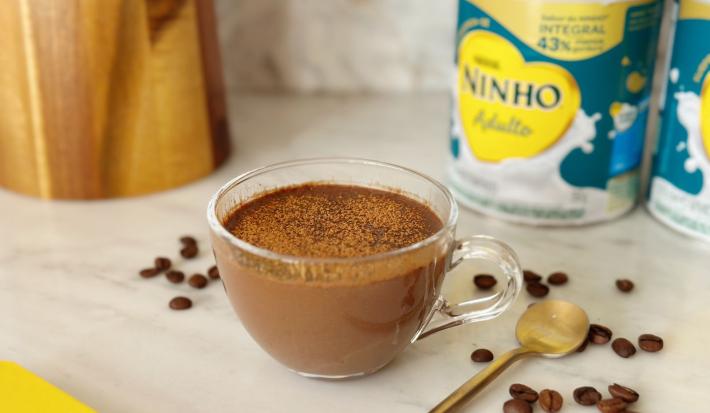 Fotografia em tons de amarelo e azul em uma bancada de mármore com uma xícara de vidro ao centro e o mocaccino dentro dela. Ao fundo, latas de Ninho Adulto e alguns grãos de café espalhados.