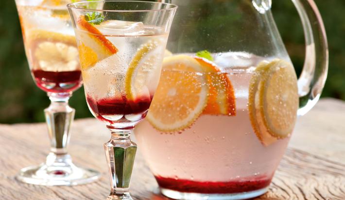 Fotografia em tons de rosa em uma bancada de madeira clara com uma jarra de vidro com o refresco de hibisco e frutas cítricas dentro, decorando com rodelas de laranja. Ao lado, duas taças de vidro com o refresco.