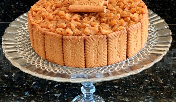 Fotografia de uma torta de biscoitos tostines, creme belga e abacaxi caramelizado. Por volta da torta tem vários tostines inteiros, e por cima, tem abacaxi em pedaços pequenos, canela em pau e mais tostines. A torta está sobre um apoio de vidro.
