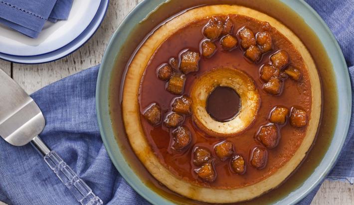 Fotografia em tons de caramelo e azul de uma mesa de madeira vista de cima, ao centro um recipiente redondo azul por cima de um tecido azul, dentro dele está o pudim com a calda de caramelo por cima e decorado com bananas carameladas.