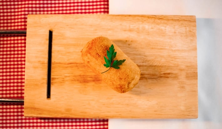 Fotografia vista de cima de um croquete frito de carne moída sobre uma tábua de madeira. Tem um pequeno ramo de salsinha decorando o croquete.