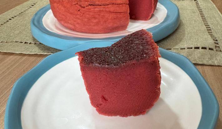 Fotografia em tons claros com um bolo mole de beterraba ao centro. O bolo tem cor rosa arroxeada e tem textura lisa e umida.
