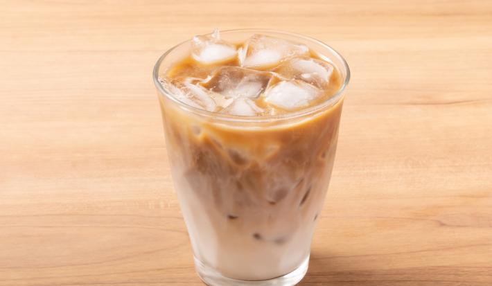 Fotografia em tons de marrom claro com um copo de vidro ao centro. Dentro do copo existe uma receita de café com leite gelado com vários cubos de gelo.