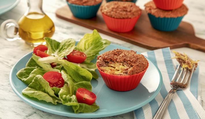 Fotografia em tons de verde em uma bancada de madeira de cor branca. Ao centro, um prato redondo azul contendo 1 muffin com um garfo ao lado. Ao fundo, uma tábua de madeira com mais muffins e um recipiente com azeite.