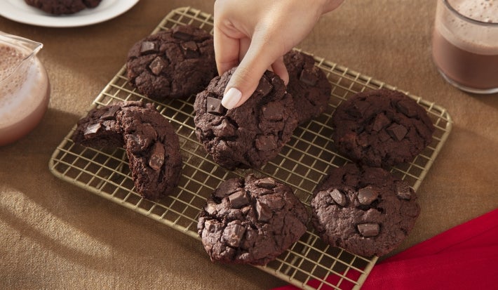 Fotografia de uma grelha quadriculada ao lado de um copo de vidro com leite e nescau. Sobre a grelha tem sete cookies de nescau extra cacau com gotas de chocolate e um mão feminina pegando um. Fotografia de uma grelha quadriculada ao lado de um copo de vidro com leite e nescau. Sobre a grelha tem sete cookies de nescau extra cacau com gotas de chocolate e um mão feminina pegando um.