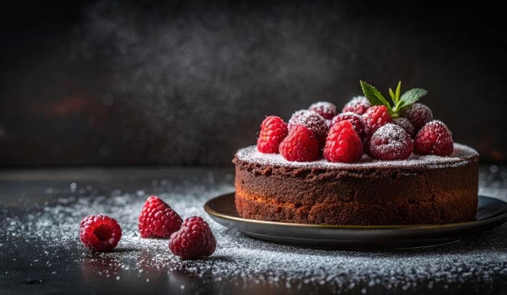 Fotografia de um bolo de chocolate rico, decorado com framboesas vermelhas e mirtilos, polvilhado com açúcar de confeiteiro. Folhas de hortelã adicionam frescor.