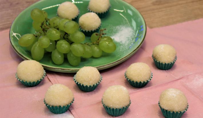Fotografia em tons de verde em uma bancada de madeira de cor marrom. Ao centro, um pano rosa contendo um prato verde em cima com os brigadeiros e um cacho de uva. Ao redor, há alguns brigadeiros espalhados.