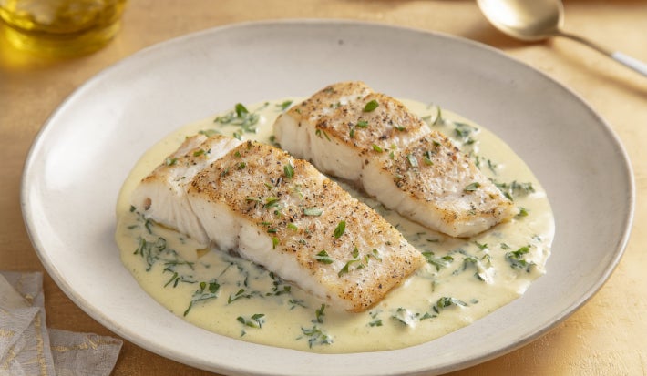 Filé de peixe branco dourado servido no molho cremoso de ervas, apresentado em prato fundo branco com acabamento sofisticado.