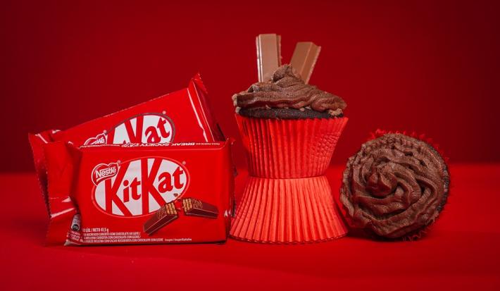 Fotografia em tons de vermelho em uma bancada de madeira de cor vermelha. Ao centro, um cupcake em pé em cima de uma forminha. AO lado direito, um cupcake deitado e ao lado esquerdo, 2 embalagens de kitkat ao laite.