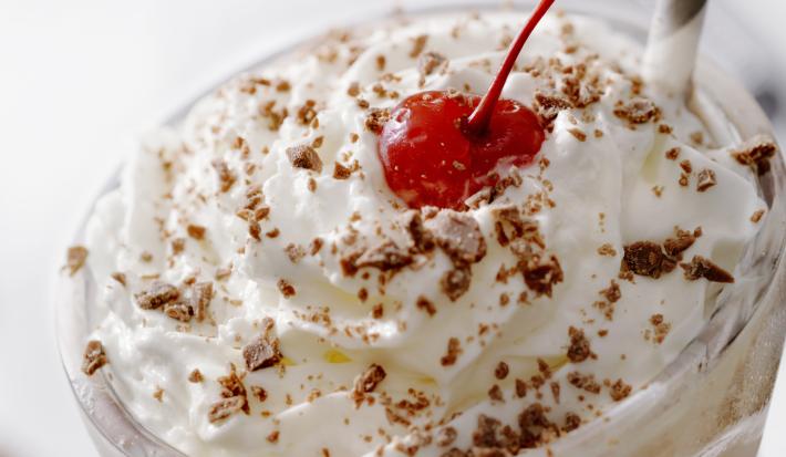 Fotografia de um copo de vidro longo com uma bebida gelada e cremosa de sorvete de chocolate, leite e chantilly. Em cima da bebida, tem raspas de chocolate e um canudo de cores alternadas e uma cereja.