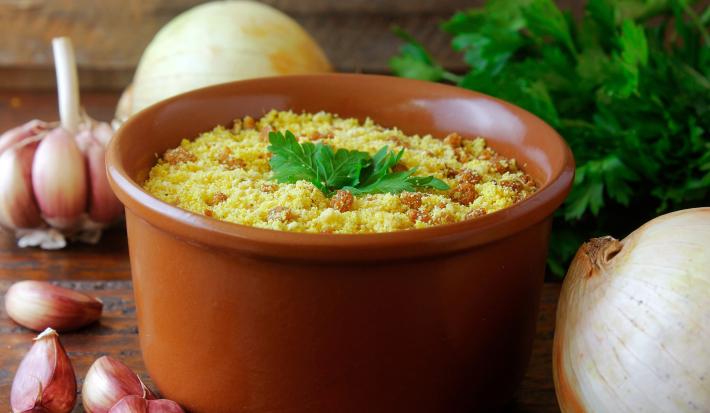 Fotografia de um recipiente fundo marrom mequeno sobre uma mesa de madeira. Dentro do recipiente tem uma farofa de pinhão com folhas de salsinha por cima, e ao redor do recipiente tem um ramo inteiro de salsinha, cebola e alho.