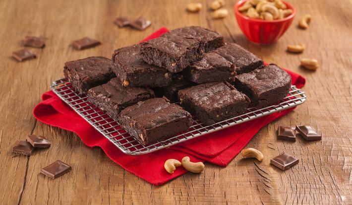 Fotografia em tons de marrom e vermelho de uma bancada de madeira com um paninho vermelho, sobre ele um prato retangular com pedaços de brownie. Ao fundo um recipiente redondo com castanha-de-caju.