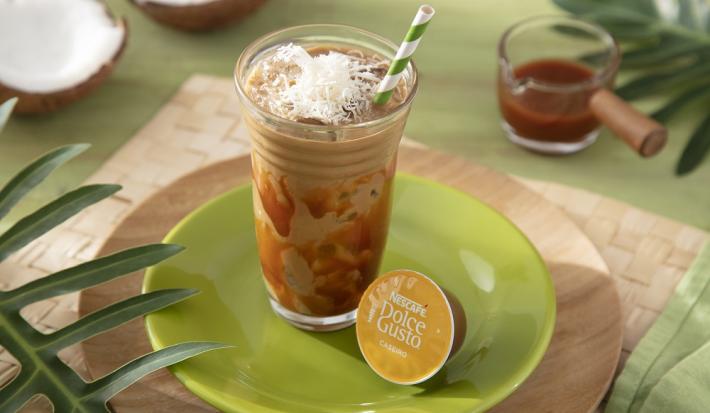 Foto da receita de iced coffee coco com caramelo servida em um copo de vidro alto com um canudo listrado verde em cima de um prato verde sobre uma tábua de madeira