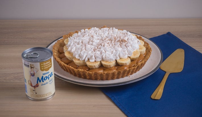 Receita de torta banoffee, servida num prato branco e decorada com chantilly e cacau em pó. A torta está ao lado de uma espátula dourada em uma bancada de madeira com um tecido na cor azul escura