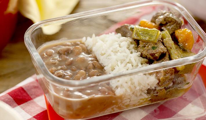 Fotografia em tons variados, com pote de vidro ao centro com feijão, arroz e picadinho de carne com legumes, sobre guardanapo quadriculado em branco e vermelho, ladeado por três panelas e uma ponta de pano amarelo ao fundo sobre bancada de madeira.