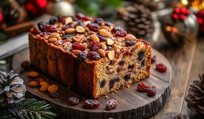 Fotografia de um bolo de frutas natalino, adornado com uma camada brilhante e coberto com nozes e frutas secas, rodeado por elementos festivos como pinhas e enfeites, evocando um clima aconchegante de celebração.