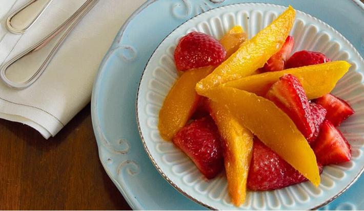 fotografia em tons de marrom, azul, amarelo e vermelho de uma bancada marrom vista de cima, contém um prato azul redondo com um pratinho redondo branco e dentro pedaços de frutas.