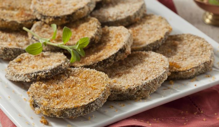 Fotografia em tons de vermelho em uma bancada de madeira com um pano vermelho, um recipiente branco retangular com as berinjelas à milanesa prontas.