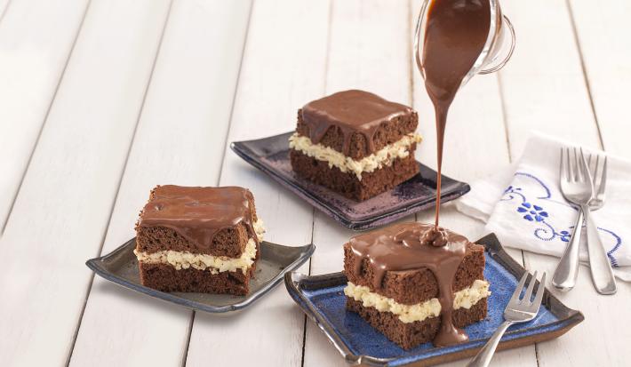 Fotografia em tons de azul em uma bancada de madeira branca, um paninho branco com detalhes azul, três pratinhos com pedaços do bolo de prestígio com calda de chocolate.