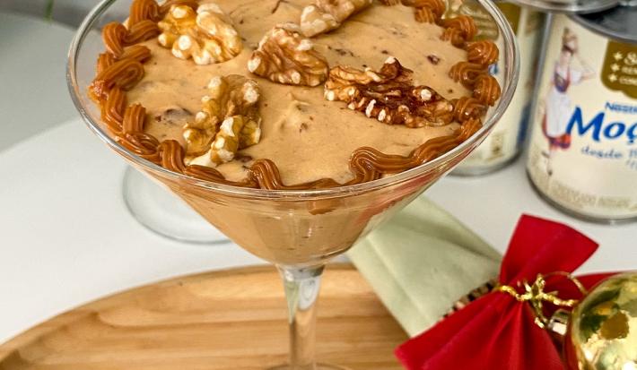 Foto em tons de bege da receita de creme natalino com nozes servida em uma taça de vidro alta sobre uma base de madeira com decoração natalina