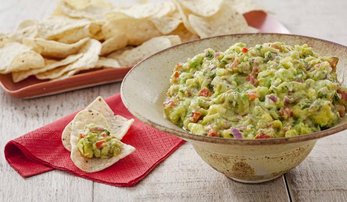 Fotografia em tons de verde e vermelho em uma bancada de madeira com um guardanapo de papel vermelho e sobre ele nachos e guacamole. Ao lado, um recipiente grande e redondo em formato de chapéu bege com o guacamole dentro dele.