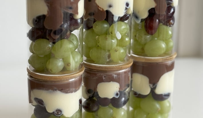 Fotografia em tons claros com vários potes ao centro. Dentro de cada pote existe uma sobremesa feita com uva, creme de leite ninho com iogurte e chocolate meio amargo