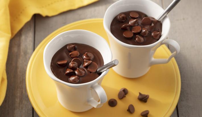 Fotografia em tons de amarelo, branco e marrom de uma bancada de madeira com um prato redondo amarelo e duas xícaras brancas com chocolate quente e uma colher. Ao fundo um paninho amarelo. Fotografia em tons de amarelo, branco e marrom de uma bancada de madeira com um prato redondo amarelo e duas xícaras brancas com chocolate quente e uma colher. Ao fundo um paninho amarelo.