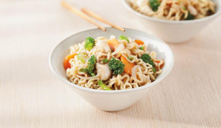 Foto de uma tigela branca com yakissoba de frango dentro e encostada nela há dois hashis. No fundo há outra tigela também com Yakissoba