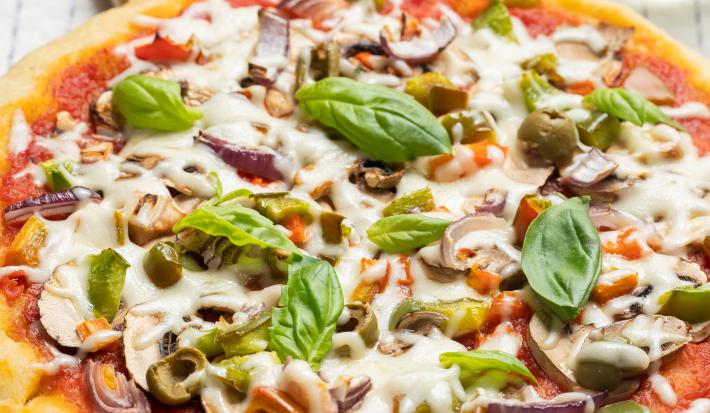 Fotografia de uma pizza com massa de pão sírio com molho de tomate, pedaços de tomate, requeijão e manjericão. A pizza está apoiada sobre uma base de madeira, que está sobre um pano de mesa branco com listras, e ao fundo, dois potinhos e um copo de vidro.