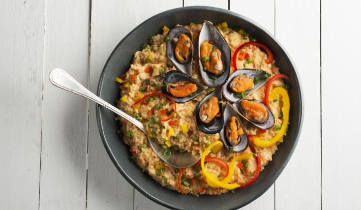 Fotografia em tons de branco e preto de uma bancada branca, sobre ele um prato preto com arroz com frutos do mar e uma colher.