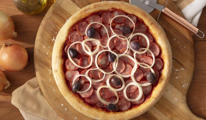 Fotografia em tons de marrom em uma bancada de madeira, uma tábua de madeira redonda com a pizza de calabresa em cima dela. Ao lado, cebola, potinho de azeite e cortador de pizza.
